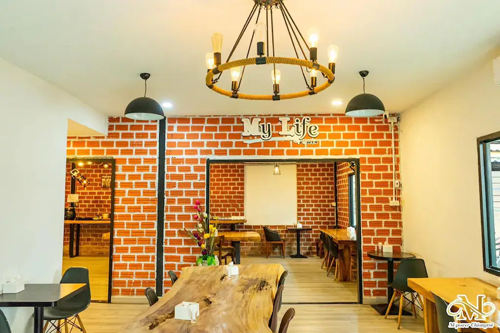 ภายในคาเฟ่ที่มีผนังลายอิฐ โต๊ะไม้ เก้าอี้สมัยใหม่ โคมไฟระย้า และป้าย "ชีวิตของฉัน" บนผนัง—เหมาะสำหรับการพักผ่อนหลังจากสำรวจ รีสอร์ทเชียงราย หรือ โรงแรมในเชียงราย.