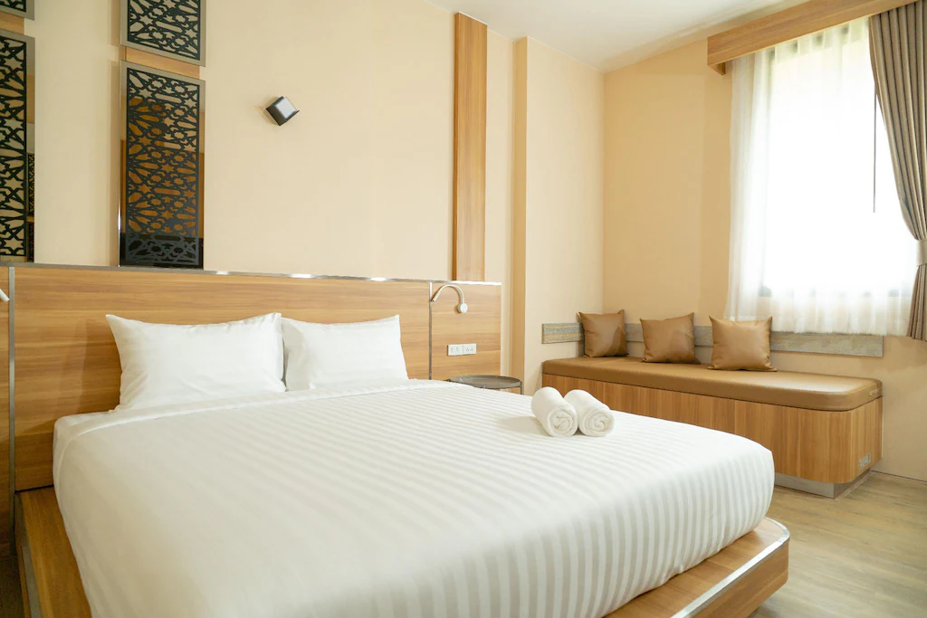 เตียงที่ปูเรียบร้อยด้วยผ้าปูที่นอนและผ้าเช็ดตัวสีขาวตั้งอยู่ในห้องนอนสไตล์โมเดิร์นโทนสว่างที่รีสอร์ทในน่าน พร้อมที่นั่งริมหน้าต่างและผ้าม่านสีเบจ.