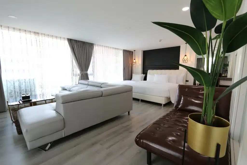 ห้องพักโรงแรมสมัยใหม่ในเชียงใหม่ ประกอบด้วยเตียงสีขาว โซฟาหนังสีเทาแบบตัวแอล ม้านั่งหนังสีน้ำตาล ต้นไม้ในกระถาง และหน้าต่างบานใหญ่จากพื้นจรดเพดานพร้อมผ้าม่านบาง.