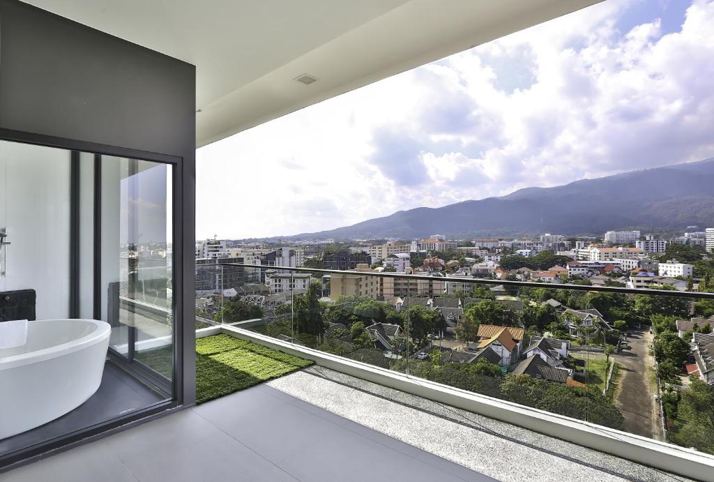 ระเบียงสมัยใหม่พร้อมราวกระจกมองเห็นทิวทัศน์เมืองและภูเขาไกล ๆ ที่โรงแรมเชียงใหม่; ห้องน้ำที่มีอ่างอาบน้ำสีขาวสามารถมองเห็นได้ผ่านประตูกระจก.
