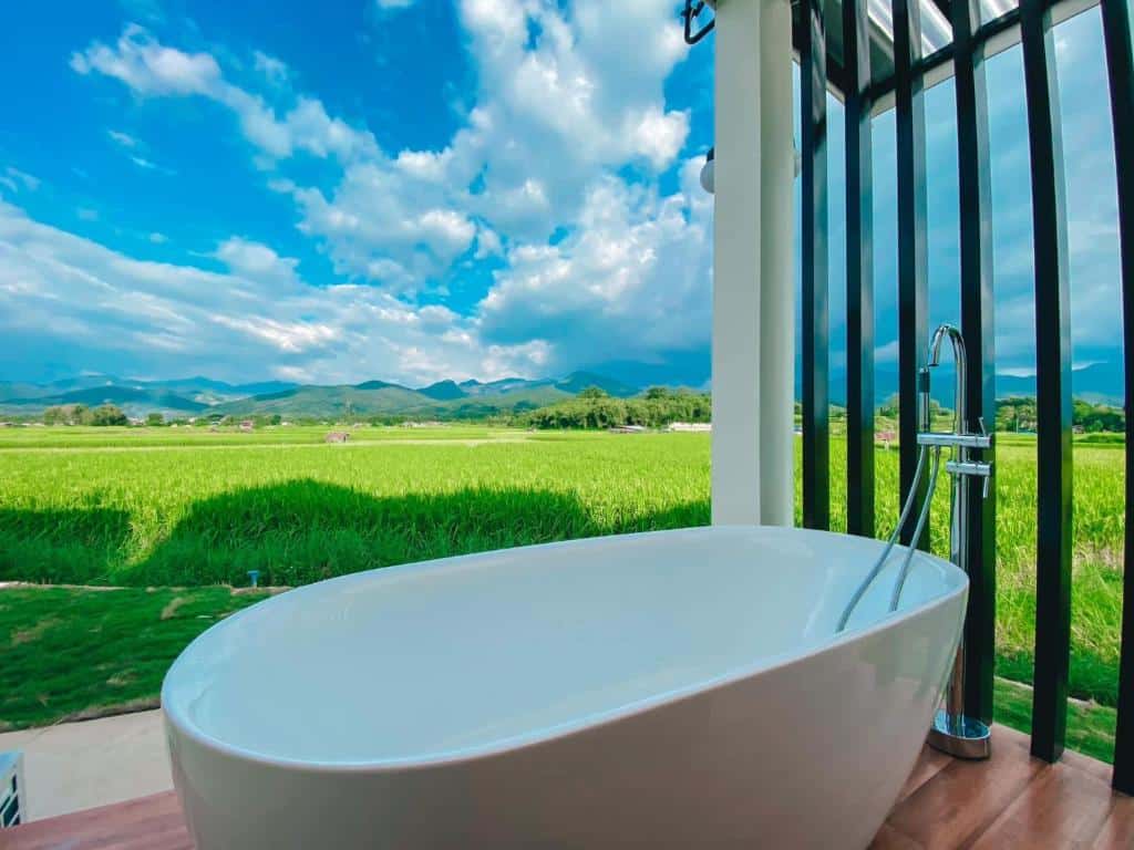 อ่างอาบน้ำสีขาวตั้งอยู่บนดาดฟ้าไม้ที่ ที่พักน่าน ให้ทัศนียภาพของทุ่งหญ้าสีเขียว ภูเขา และท้องฟ้าที่มีเมฆบางส่วนเป็นฉากหลัง.