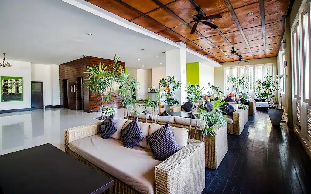 พื้นที่เลานจ์สมัยใหม่ที่โรงแรมเชียงใหม่ มีโซฟาหวาย, หมอนสีเทา, ต้นไม้ในกระถาง, พื้นไม้สีเข้ม, และเพดานไม้พร้อมพัดลมเพดาน แสงธรรมชาติส่องผ่านหน้าต่างบานใหญ่.
