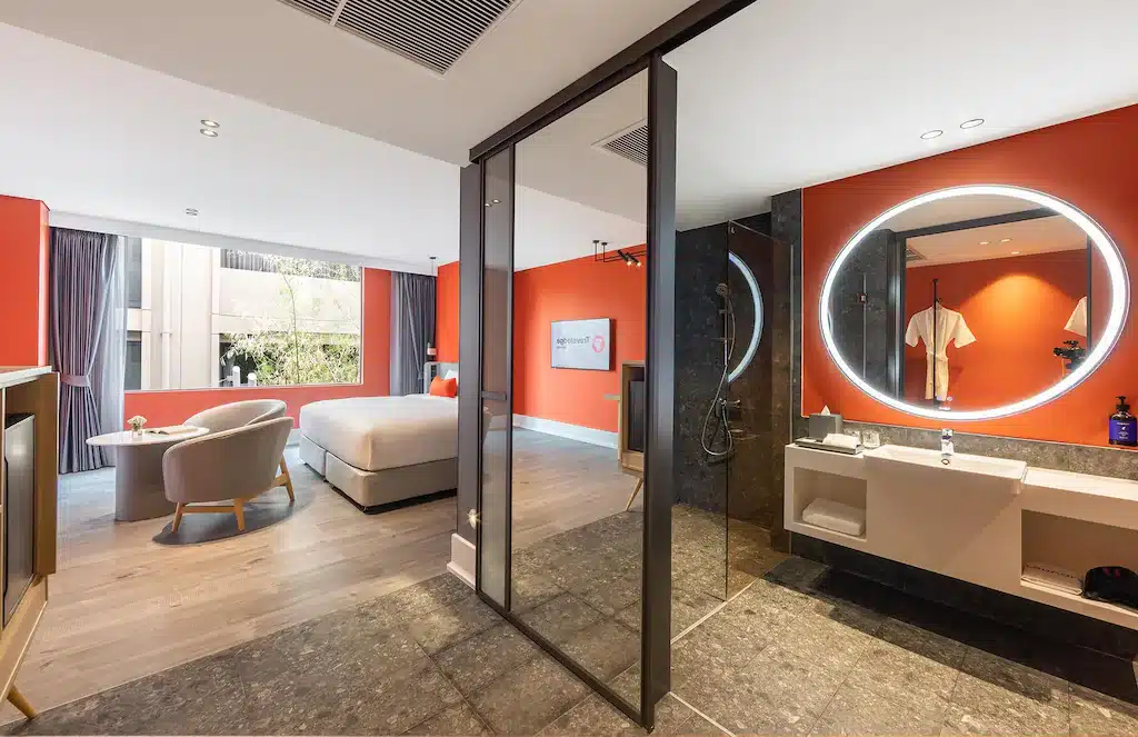 ห้องพักโรงแรมสมัยใหม่ที่ โรงแรมเชียงใหม่ แห่งนี้มีเตียงขนาดใหญ่, บริเวณนั่งเล่น, และห้องน้ำแบบเปิดโล่งพร้อมกระจกกลมส่องสว่างและฝักบัวแบบกระจก ผนังสีส้มและสีกลาง ๆ ช่วยเสริมให้กับการออกแบบที่กว้างขวาง หน้าต่างขนาดใหญ่ช่วยให้แสงธรรมชาติส่องเข้ามา.