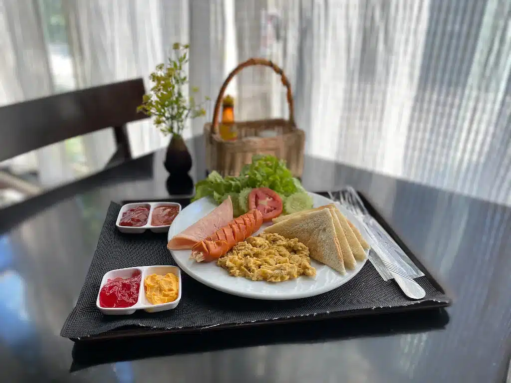 จานอาหารเช้าประกอบด้วยไข่คน ไส้กรอก ขนมปังปิ้ง มะเขือเทศ ผักกาดหอม และเครื่องปรุงรสต่างๆ บนถาดสีดำ ที่รีสอร์ทเชียงใหม่ พร้อมตะกร้าและแจกันดอกไม้เล็กๆ บนโต๊ะ.