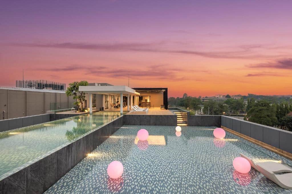 สระว่ายน้ำอินฟินิตี้บนดาดฟ้าพร้อมไฟสีชมพูลอยน้ำยามพระอาทิตย์ตกดิน ติดกับพื้นที่เลานจ์ทันสมัยที่มีต้นไม้และวิวเมือง—เหมาะสำหรับการพักผ่อนที่ โรงแรมเชียงใหม่ ที่มีสไตล์.