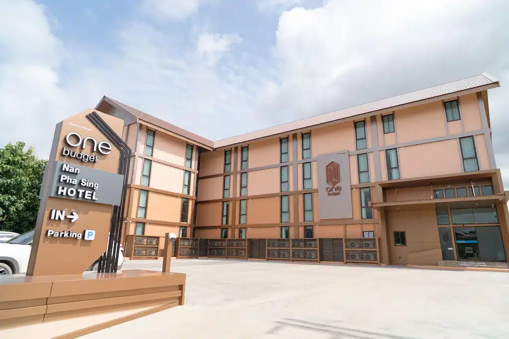 โรงแรมสมัยใหม่สามชั้น ตกแต่งภายนอกด้วยโทนสีน้ำตาลและสีแทน มีป้ายชื่อโรงแรมขนาดใหญ่ที่อ่านว่า "โรงแรมวัน บัดเจ็ต น่าน พะสิงห์" พร้อมลานจอดรถด้านหน้าภายใต้ท้องฟ้าที่มีเมฆบางส่วน—เหมาะสำหรับผู้ที่มองหาที่พักในน่านหรือโรงแรมในน่าน.