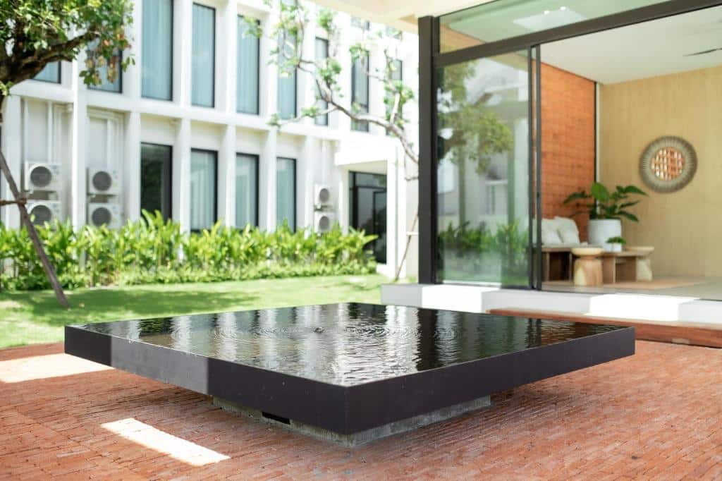 น้ำพุสะท้อนแสงสีดำสมัยใหม่ตั้งอยู่บนลานอิฐในลานกลางแจ้งที่ตกแต่งอย่างสวยงามของรีสอร์ทเชียงใหม่สไตล์หรูแห่งนี้ ติดกับห้องผนังกระจกที่มีที่นั่งและต้นไม้ในร่ม.