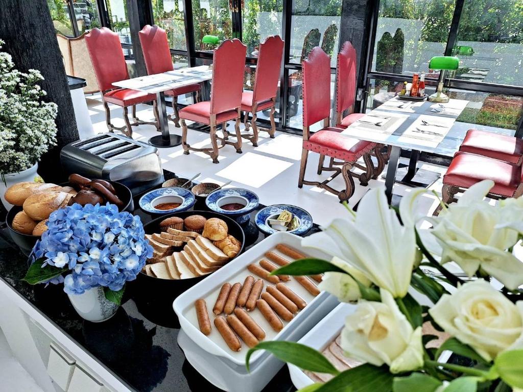 บุฟเฟ่ต์อาหารเช้าประกอบด้วยขนมปัง ไส้กรอก ขนมอบ และเครื่องเคียงต่างๆ จัดวางอยู่บนเคาน์เตอร์ที่ โรงแรมในเชียงราย โดยมีโต๊ะและเก้าอี้สีแดงจัดเรียงอยู่ในพื้นที่รับประทานอาหารที่สว่างสดใสในพื้นหลัง.