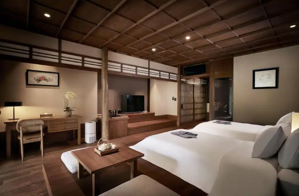 ห้องพักสไตล์ญี่ปุ่นสมัยใหม่ในโรงแรมเชียงใหม่ ประกอบด้วยเตียงสองเตียง โต๊ะไม้ โทรทัศน์ โต๊ะต่ำ แสงสว่างนุ่มนวล ตกแต่งแบบดั้งเดิม และผนังไม้ทั่วทั้งห้อง—เหมาะสำหรับการเข้าพักของคุณในเชียงใหม่.