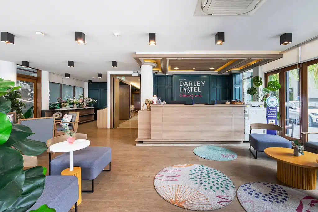 ล็อบบี้โรงแรมสมัยใหม่พร้อมพื้นไม้ เฟอร์นิเจอร์ร่วมสมัย และพรมวงกลมสีสันสดใส ต้อนรับแขกสู่โรงแรมดาร์ลีย์ เชียงใหม่ หน้าต่างบานใหญ่เปิดรับแสงธรรมชาติเข้าสู่พื้นที่—ทางเลือกที่มีสไตล์สำหรับโรงแรมในเชียงใหม่หรือที่พักในเชียงใหม่.
