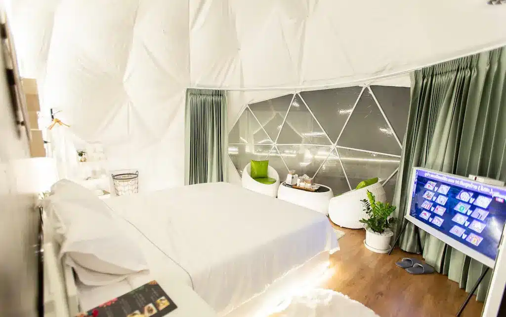 ห้องนอนทรงโดมจีโอเดสิกสมัยใหม่ที่ ที่พักเชียงใหม่ โดดเด่นด้วยเตียงคู่ ทีวี เก้าอี้สองตัวริมหน้าต่างบานใหญ่พร้อมผ้าม่านสีเขียว พื้นไม้ และการตกแต่งแบบมินิมอล เหมาะสำหรับการพักผ่อนในเชียงใหม่อย่างสมบูรณ์แบบ.