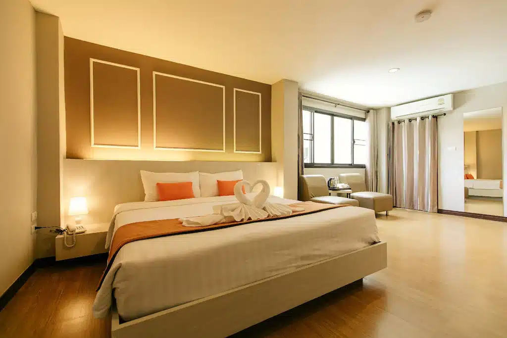 ห้องพักโรงแรมเชียงรายสไตล์โมเดิร์นพร้อมเตียงขนาดใหญ่, รายละเอียดสีส้ม, นกผ้าขนหนู, โคมไฟข้างเตียง, มุมนั่งเล่นริมหน้าต่าง, และกระจกติดผนัง.