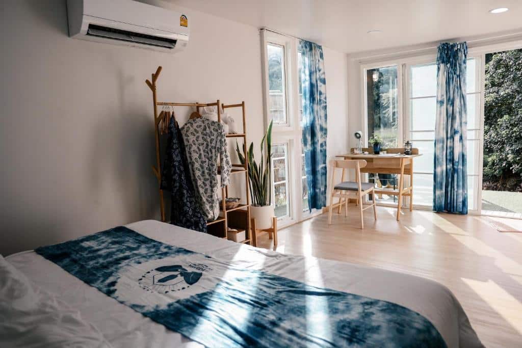 ห้องนอนสว่างสไตล์มินิมอลพร้อมชุดเครื่องนอนลายมัดย้อมสีน้ำเงิน โต๊ะไม้ข้างหน้าต่างบานใหญ่ ต้นไม้架เสื้อผ้า และเครื่องปรับอากาศ—เหมาะสำหรับนักเดินทางที่กำลังมองหาที่พักในน่านหรือโรงแรมในน่านในบรรยากาศที่มีสไตล์และสะดวกสบาย.