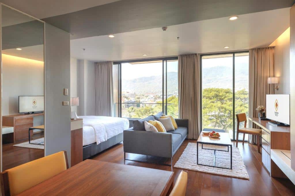 ห้องสวีทโรงแรมสมัยใหม่ที่ โรงแรมเชียงใหม่ พร้อมเตียง โซฟา โต๊ะ โทรทัศน์ และหน้าต่างบานใหญ่ที่มองเห็นวิวต้นไม้และภูเขาภายนอก.