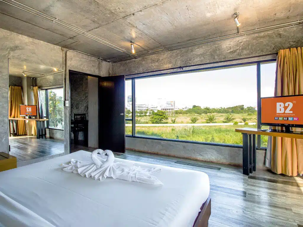 ห้องพักโรงแรมสมัยใหม่พร้อมผนังคอนกรีต เตียงขนาดใหญ่ประดับด้วยหงส์ผ้าขนหนู หน้าต่างบานกว้างมองเห็นวิวสีเขียวสดใส ทีวีสองเครื่อง และกระจกที่สะท้อนส่วนหนึ่งของห้อง—เหมาะสำหรับผู้ที่มองหาที่พักเชียงใหม่ที่ไม่เหมือนใคร.