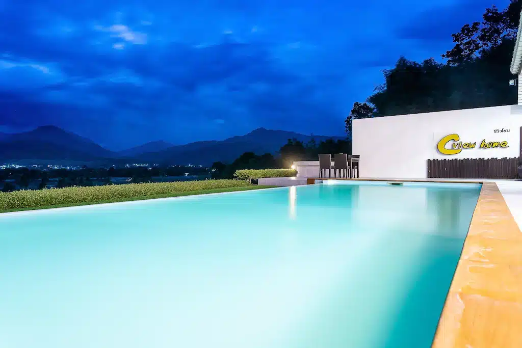 สระว่ายน้ำไร้ขอบพร้อมน้ำสีฟ้าเรืองแสงยามพลบค่ำ มองเห็นทิวทัศน์อันงดงามของภูเขาและแสงไฟในระยะไกลใต้ท้องฟ้าที่มีเมฆปกคลุม ที่รีสอร์ทในน่าน.