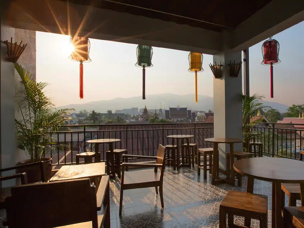 ระเบียงดาดฟ้าที่รีสอร์ทเชียงใหม่แห่งหนึ่งมีโต๊ะและเก้าอี้ไม้ตั้งอยู่ มองเห็นวิวเส้นขอบฟ้าของเมืองยามพระอาทิตย์ตกดิน พร้อมโคมไฟสีสันสดใสแขวนอยู่ด้านบนและภูเขาเป็นฉากหลัง.