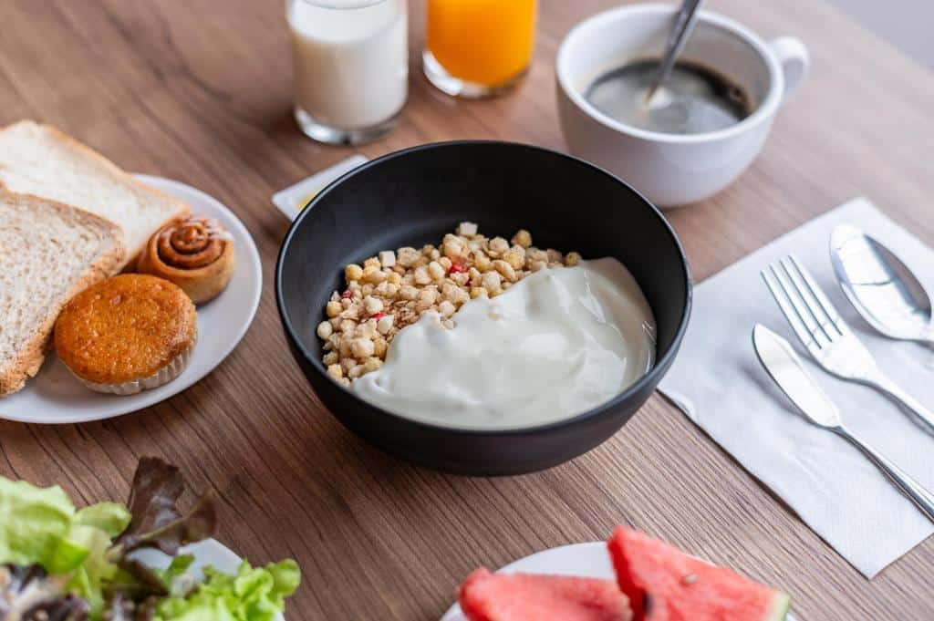 ชามกราโนล่าพร้อมโยเกิร์ต ขนมปังปิ้ง ขนมอบ คัพเค้ก สลัด แตงโม นม น้ำส้ม และกาแฟ บนโต๊ะไม้—เหมาะสำหรับการเริ่มต้นเช้าวันใหม่ที่รีสอร์ทแสนอบอุ่นในเชียงราย.