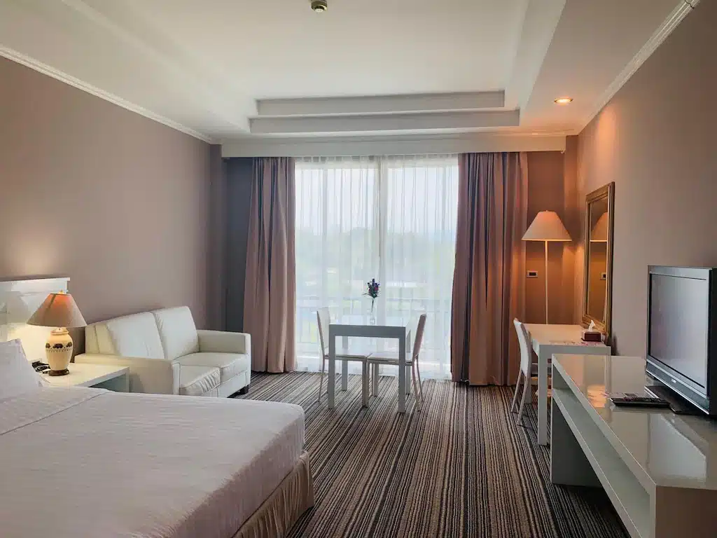 ห้องพักโรงแรมที่แสนสบายที่ โรงแรมเชียงราย พร้อมเตียงนอนที่นุ่มสบาย โซฟาขาว โต๊ะอาหารเล็กสำหรับสองคน โต๊ะทำงาน โทรทัศน์ และหน้าต่างบานใหญ่พร้อมผ้าม่านที่เปิดให้แสงสว่างเข้ามา.