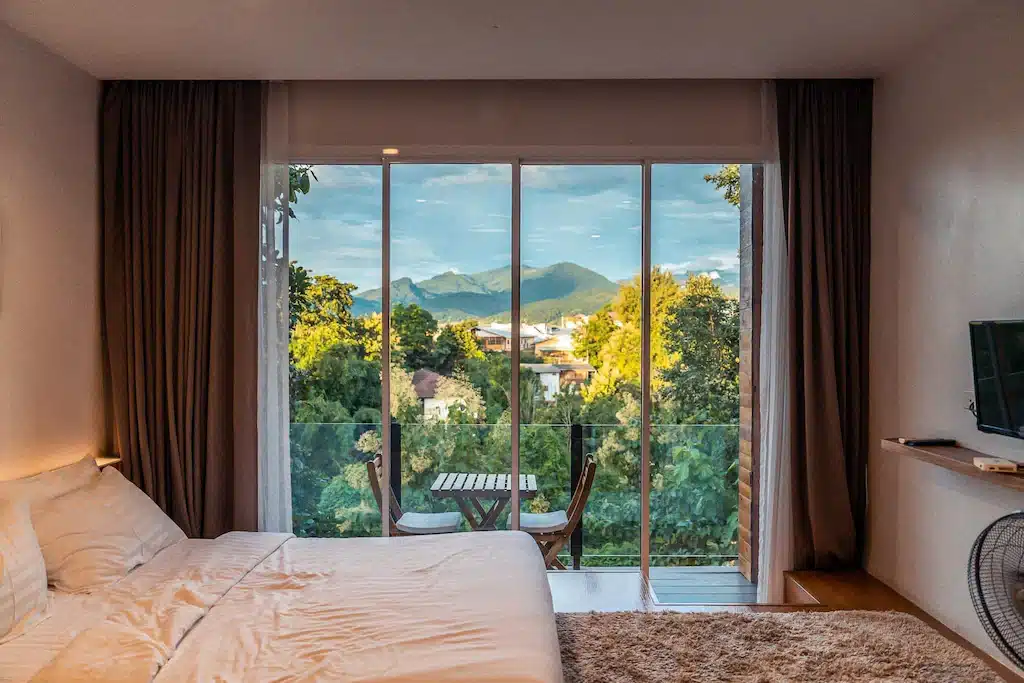 เตียงที่จัดไว้อย่างเรียบร้อยหันหน้าไปทางประตูกระจกที่เปิดออกสู่ระเบียงซึ่งมีโต๊ะและเก้าอี้สองตัว มองเห็นต้นไม้และภูเขาไกลๆ ภายใต้ท้องฟ้าสีคราม ณ ที่พักแสนมีเสน่ห์แห่งนี้ในน่าน.