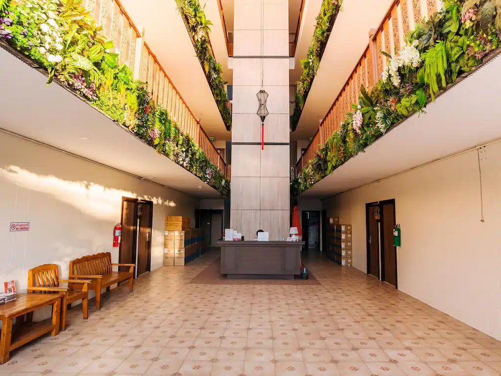 ล็อบบี้ที่กว้างขวางพร้อมพื้นกระเบื้อง โต๊ะต้อนรับตรงกลาง ม้านั่งไม้ทางด้านซ้าย และทางเดินชั้นบนสองทางประดับด้วยต้นไม้สีเขียว ต้อนรับคุณสู่โรงแรมสุดมีสไตล์ในเชียงใหม่แห่งนี้.