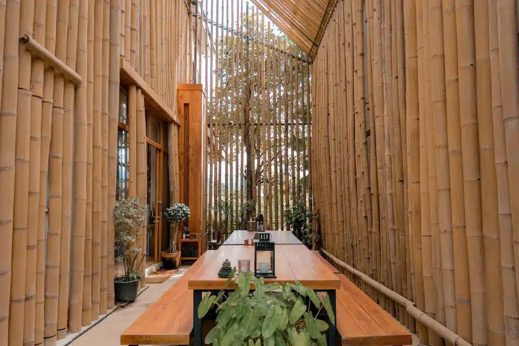 โต๊ะอาหารไม้ยาวพร้อมต้นไม้ในกระถางตั้งอยู่ในห้องแคบสูงที่มีผนังและเพดานทำจากไม้ไผ่ตั้งตรงที่รีสอร์ทน่ารักในน่าน ต้นไม้สามารถมองเห็นได้ผ่านช่องไม้ไผ่ สร้างบรรยากาศที่สงบเงียบ.