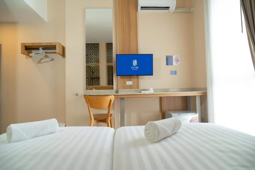 ห้องพักโรงแรมพร้อมเตียงสองเตียง โทรทัศน์ติดผนัง โต๊ะพร้อมเก้าอี้ กระจก ตู้เย็นขนาดเล็ก และผ้าเช็ดตัวพับไว้บนเตียง—เหมาะสำหรับการเข้าพักของคุณที่ ที่พักน่าน หรือ รีสอร์ทน่าน.