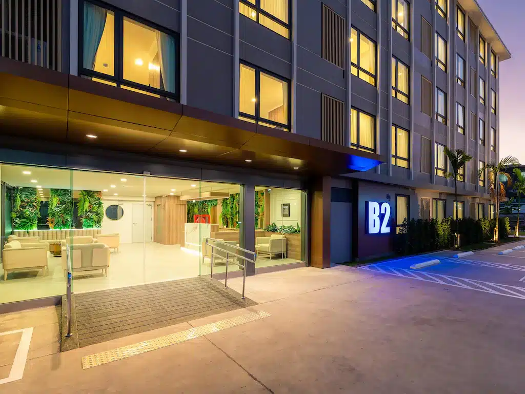 ภายนอกของโรงแรมเชียงใหม่สมัยใหม่ที่มีหน้าต่างบานใหญ่, ป้ายไฟ "B2", ทางเข้าที่เป็นกระจก, ทางลาดสำหรับผู้พิการ, และที่จอดรถว่างในยามพลบค่ำ.