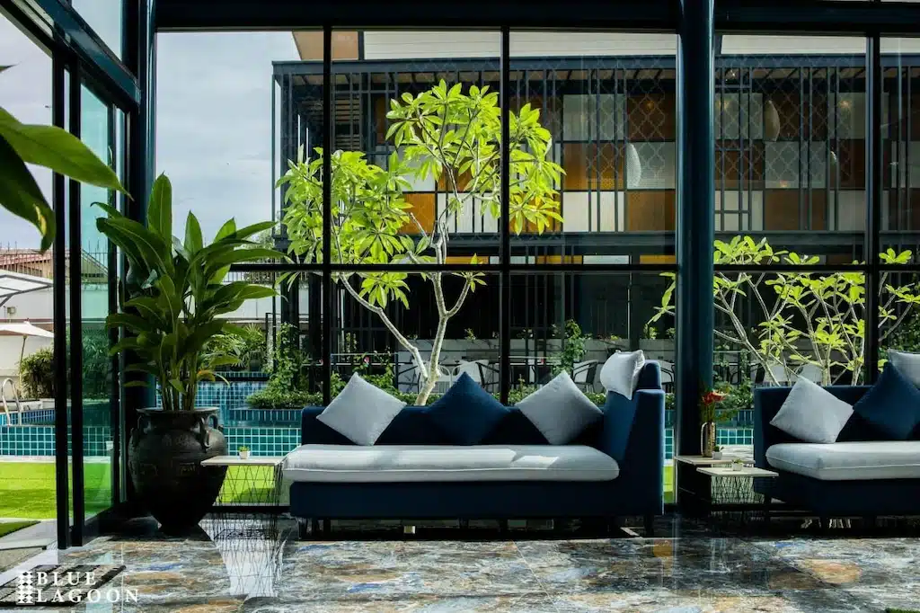 พื้นที่เลานจ์สมัยใหม่ที่ โรงแรมเชียงราย พร้อมโซฟาสีน้ำเงิน หมอนประดับ และหน้าต่างบานใหญ่ที่มองเห็นสระว่ายน้ำและสวนเขียวชอุ่ม.