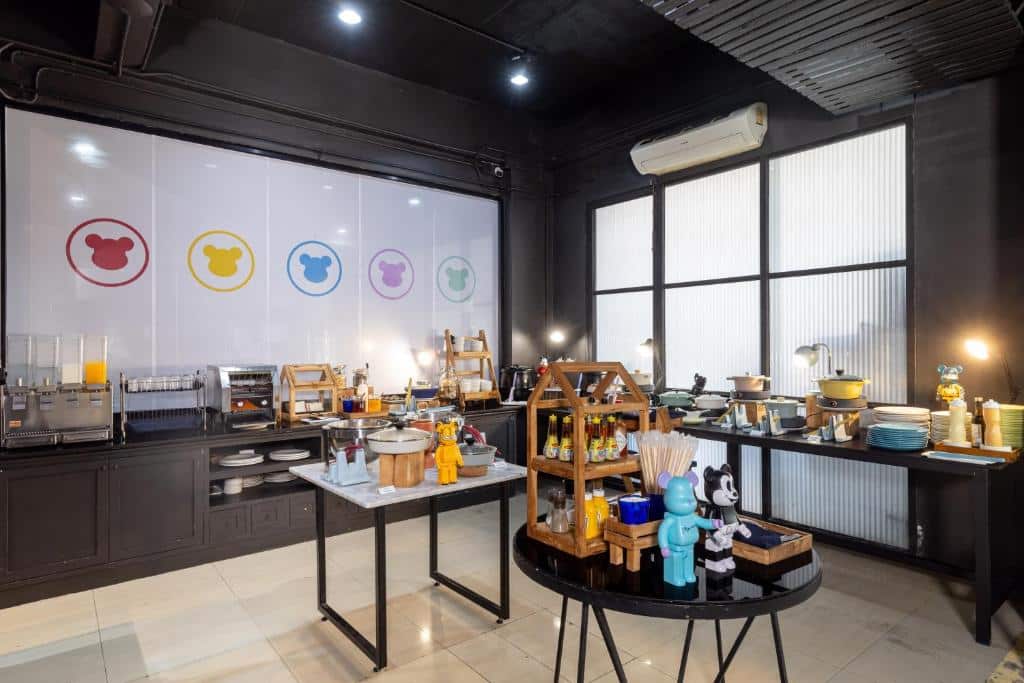 การจัดบุฟเฟ่ต์ในห้องที่มีแสงสว่างเพียงพอ พร้อมเครื่องดื่ม เครื่องปรุง เครื่องใช้ และอาหารต่างๆ บนโต๊ะ; ไอคอนรูปหมีการ์ตูนประดับผนังด้านหลัง สร้างบรรยากาศที่ร่าเริงและเหมาะสมกับรีสอร์ทเชียงใหม่ที่เป็นมิตรกับครอบครัว.