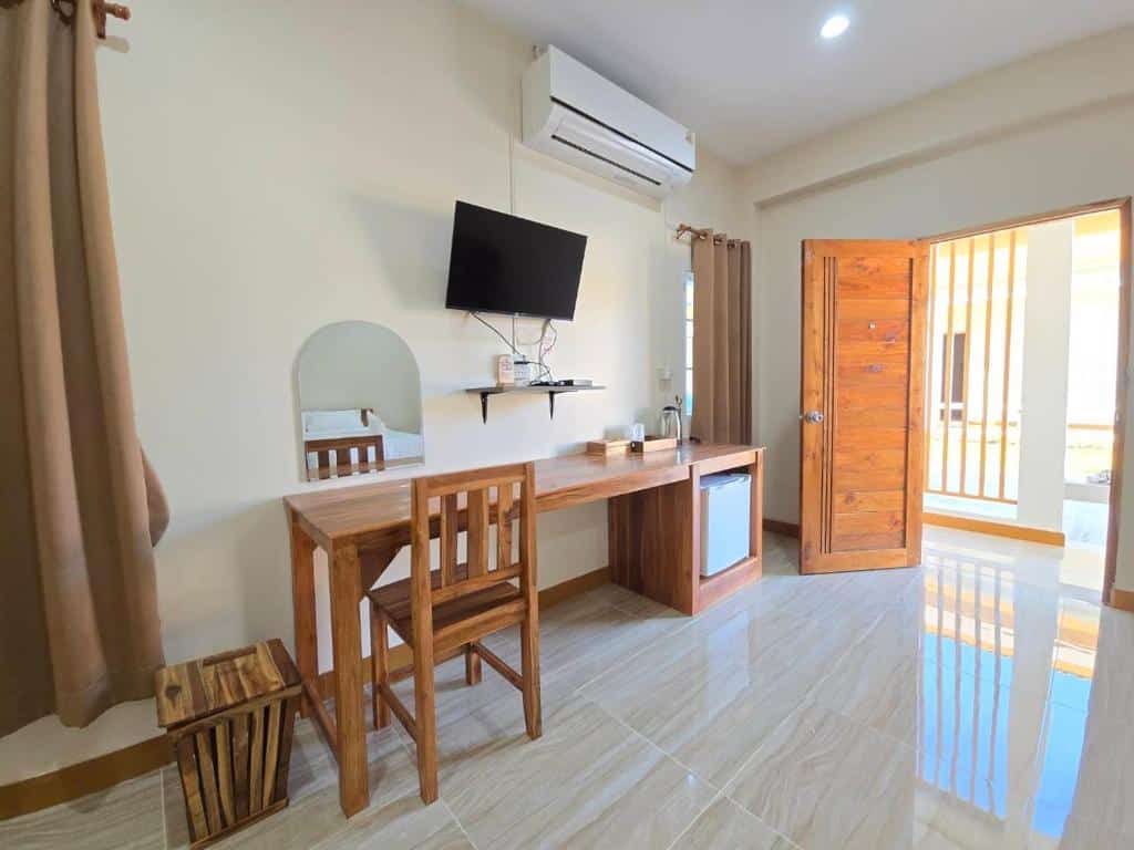 ห้องพักเรียบง่ายที่รีสอร์ทนาน พร้อมโต๊ะและเก้าอี้ไม้ โทรทัศน์ติดผนัง ตู้เย็นขนาดเล็ก เครื่องปรับอากาศ และประตูไม้เปิดโล่งรับแสงธรรมชาติ เหมาะสำหรับการพักผ่อนที่ที่พักนานของคุณ.