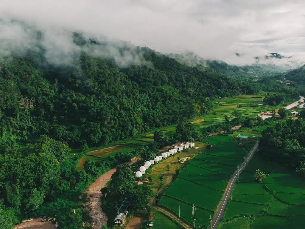 ภาพมุมสูงของทุ่งข้าวสีเขียว แม่น้ำ และแถวเต็นท์สีขาวใกล้ภูเขาที่ปกคลุมด้วยป่าและเมฆต่ำ—รีสอร์ทในฝันที่ซ่อนตัวอยู่ในธรรมชาติของน่าน.