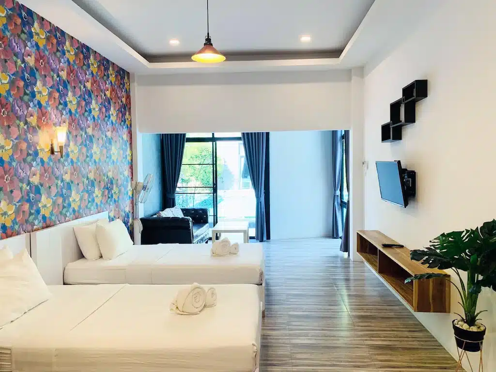 ห้องพักโรงแรมสมัยใหม่ที่ โรงแรมน่าน พร้อมเตียงเดี่ยวสองเตียง ผนังตกแต่งด้วยดอกไม้สีสันสดใส ทีวีติดผนัง พื้นไม้ โซฟาใกล้หน้าต่าง และต้นไม้ในกระถางที่มุมห้อง.