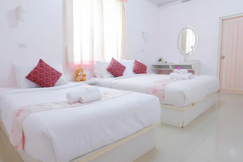 ห้องนอนที่สว่างและเรียบร้อยที่รีสอร์ทในน่าน พร้อมเตียงเดี่ยวสองเตียง หมอนลายสีชมพู ผ้าปูที่นอนสีขาว โต๊ะเครื่องแป้งขนาดเล็ก หน้าต่างพร้อมผ้าม่านบาง และตุ๊กตาผ้าบนเตียงหนึ่งเตียง—เหมาะอย่างยิ่งสำหรับประสบการณ์การพักผ่อนในน่านของคุณ.