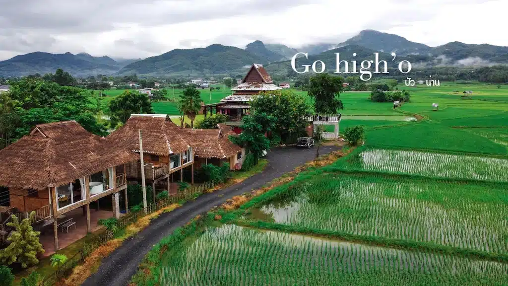 ทิวทัศน์ชนบทที่มีบ้านหลังคามุงจาก รีสอร์ทในน่านล้อมรอบด้วยทุ่งนาสีเขียวขจี ภูเขาเป็นฉากหลัง และข้อความ "Go high'o" ในท้องฟ้า.