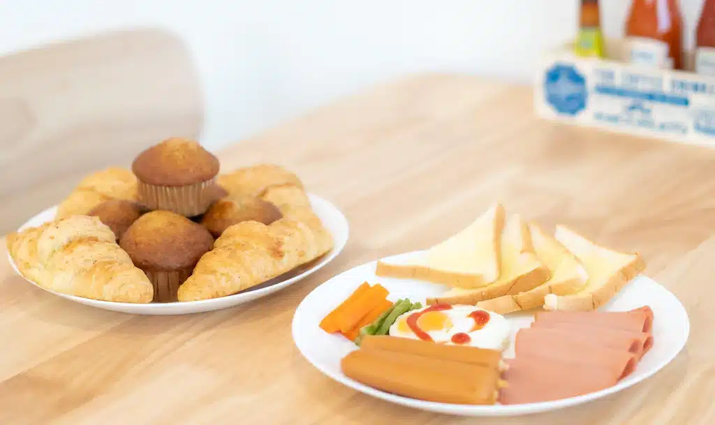 จานสองใบบนโต๊ะไม้ที่รีสอร์ทเชียงราย: ใบหนึ่งมีครัวซองต์และมัฟฟิน อีกใบมีขนมปังปิ้ง ไส้กรอกหั่นเป็นชิ้น แครอทแท่ง ถั่วแขก และไข่ดาวราดซอสมะเขือเทศ.