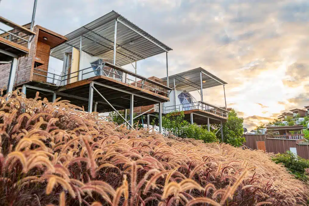 บ้านสมัยใหม่ยกสูงที่มีหลังคาโลหะและหน้าต่างบานใหญ่ถูกสร้างขึ้นบนเสาสูงเหนือทุ่งหญ้าสีน้ำตาลแดงสูงใหญ่ ดูคล้ายที่พักเชียงใหม่ที่มีเอกลักษณ์เฉพาะตัว ท่ามกลางท้องฟ้ายามพระอาทิตย์ตกที่เต็มไปด้วยเมฆ.