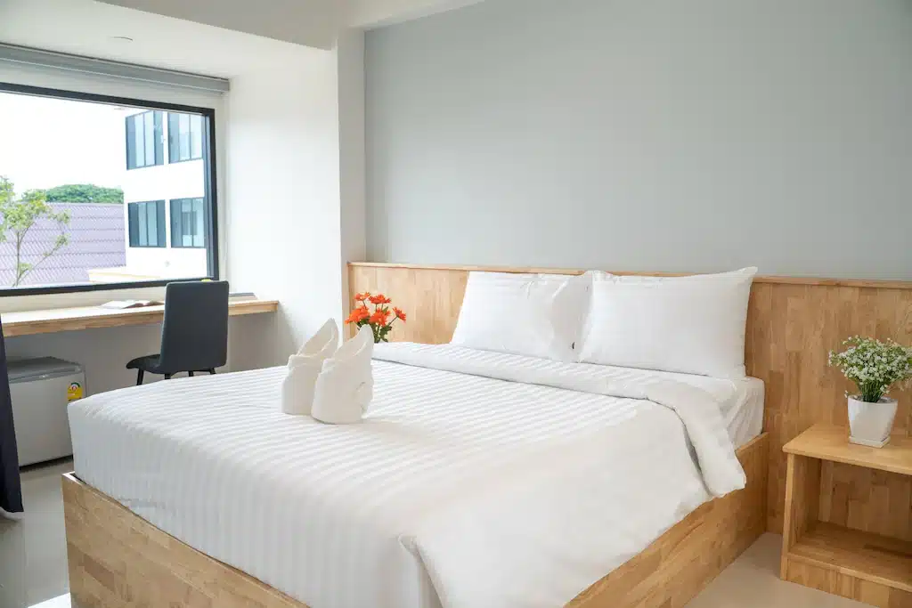 ห้องนอนสมัยใหม่ที่โรงแรมเชียงราย พร้อมเตียงขนาดใหญ่ ผ้าปูที่นอนสีขาว ผ้าเช็ดตัวรูปหงส์ โต๊ะข้างเตียงไม้พร้อมดอกไม้ โต๊ะทำงาน เก้าอี้สำนักงาน ตู้เย็นขนาดเล็ก และหน้าต่างบานใหญ่ที่เปิดรับแสงธรรมชาติ.