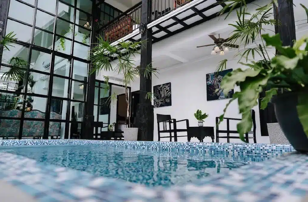 สระว่ายน้ำในร่มพร้อมกระเบื้องสีฟ้าล้อมรอบ ผนังกระจกกรอบสีดำ ต้นไม้ในกระถาง เก้าอี้ และพัดลมเพดานในพื้นที่เปิดโล่งทันสมัยที่ โรงแรมเชียงราย ที่มีสไตล์.