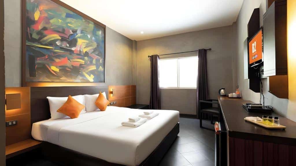 ห้องพักโรงแรมสมัยใหม่ที่ โรงแรมเชียงใหม่ พร้อมเตียงคู่ หมอนสีส้มสองใบ ภาพวาดนามธรรมขนาดใหญ่เหนือหัวเตียง โทรทัศน์ติดผนัง โต๊ะทำงาน และหน้าต่างพร้อมม่านปิด.