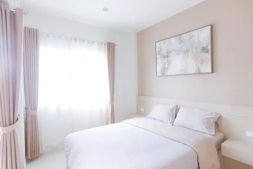 ห้องนอนสไตล์มินิมอลที่ โรงแรมเชียงราย ประกอบด้วยเตียงคู่ที่จัดไว้อย่างเรียบร้อย ผ้าม่านสีอ่อนที่ล้อมกรอบหน้าต่างสว่าง และภาพวาดนามธรรมเหนือหัวเตียง.