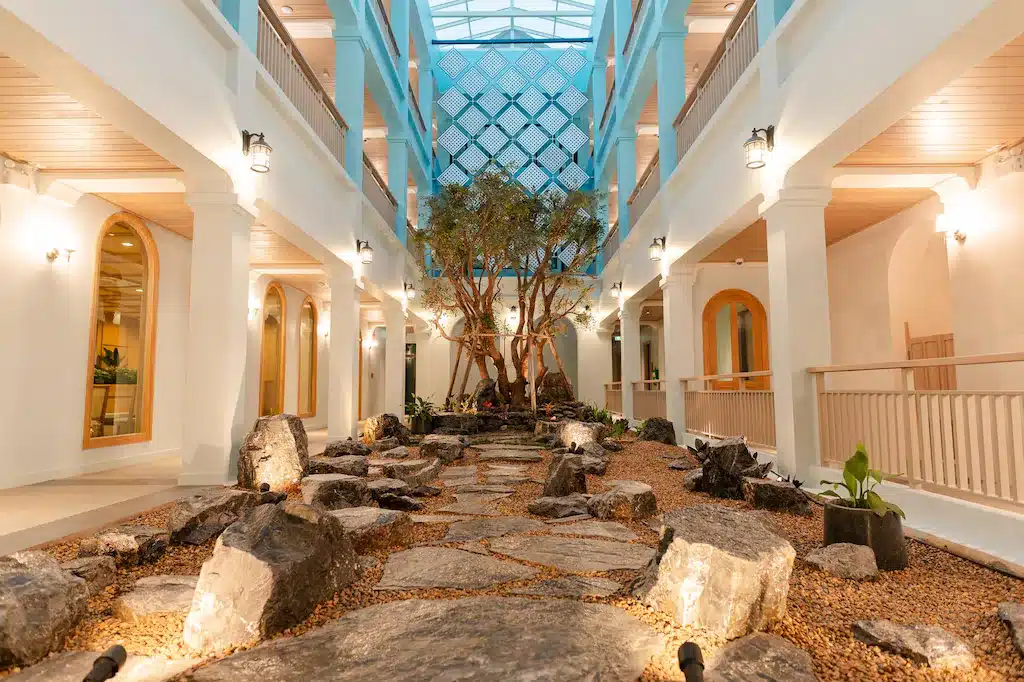 ลานภายในที่มีหลังคากระจก พร้อมทางเดินหิน หินธรรมชาติ พืชขนาดเล็ก และต้นไม้ใหญ่ตรงกลาง—เหมาะสำหรับการพักผ่อนที่ โรงแรมน่าน—ล้อมรอบด้วยเสาหินสีขาวและราวไม้.