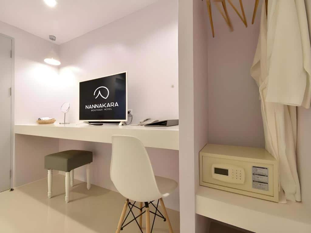 ห้องพักสไตล์มินิมอลที่โรงแรมน่าน พร้อมโต๊ะสีขาว เก้าอี้ ทีวีแสดงข้อความ "NANNAKARA Boutique Hotel" เก้าอี้สตูล โคมไฟติดผนัง ตู้เซฟ และเสื้อคลุมสีขาวแขวนอยู่ในตู้เสื้อผ้าแบบเปิด—เหมาะอย่างยิ่งสำหรับประสบการณ์การพักผ่อนในน่านครั้งต่อไปของคุณ.