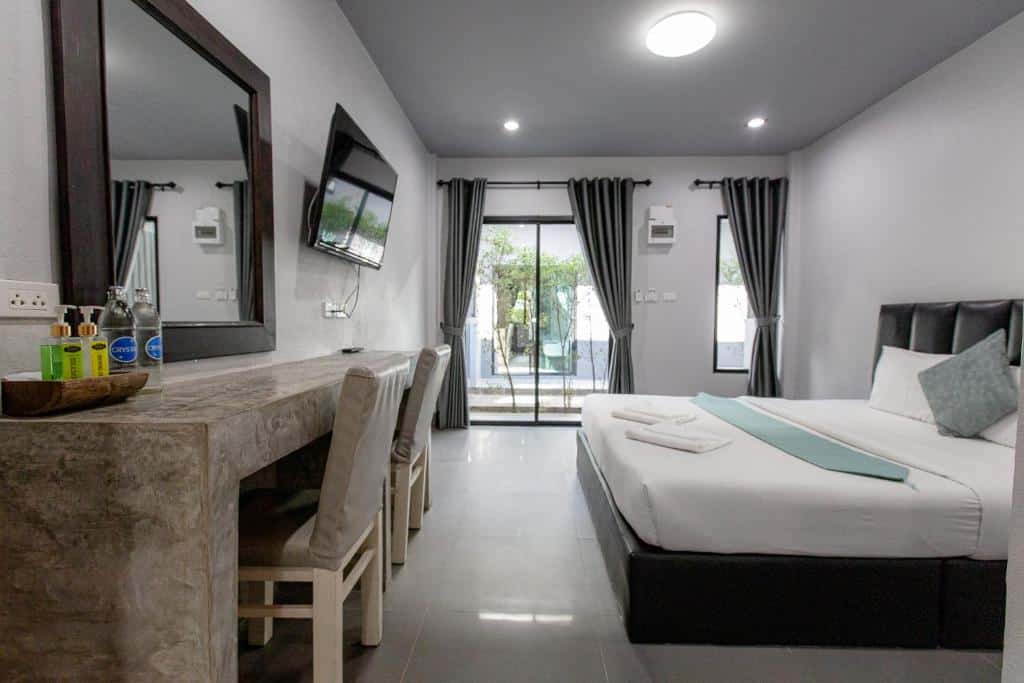 ห้องพักโรงแรมสมัยใหม่ที่ ที่พักเชียงราย พร้อมเตียงขนาดใหญ่ ทีวีติดผนัง โต๊ะคอนกรีตและเก้าอี้ เครื่องใช้ในห้องน้ำ และหน้าต่างพร้อมผ้าม่านที่เปิดออกสู่พื้นที่ลานด้านนอก.