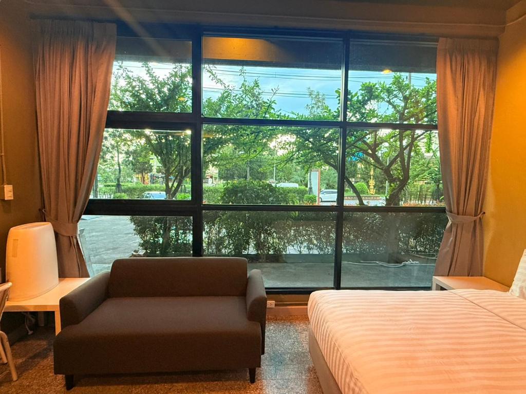 ห้องพักแสนสบายที่รีสอร์ทเชียงราย พร้อมเตียงนอน โซฟาขนาดเล็กสีเข้ม โต๊ะทำงาน และหน้าต่างบานใหญ่พร้อมผ้าม่าน มองเห็นต้นไม้และรถยนต์ที่จอดอยู่ด้านนอก.