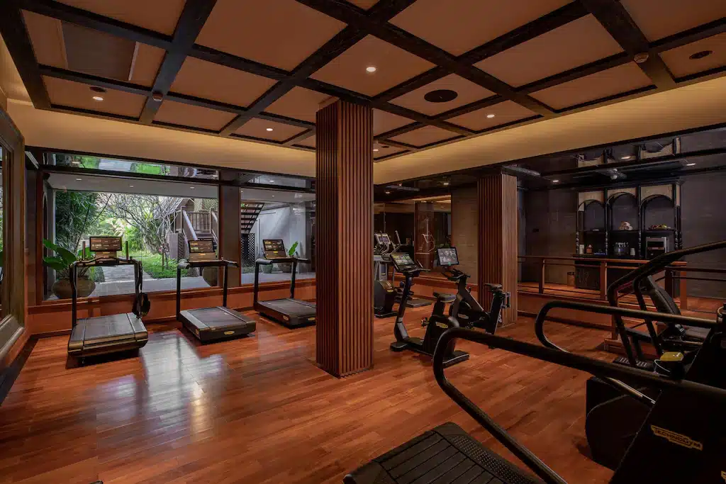 ห้องออกกำลังกายสมัยใหม่ที่รีสอร์ทเชียงใหม่ของเรา มีพื้นไม้ เครื่องวิ่งออกกำลังกาย จักรยานออกกำลังกาย และหน้าต่างขนาดใหญ่ที่มองเห็นสวนได้ แสงไฟที่อบอุ่นและแผ่นเพดานสร้างบรรยากาศที่แสนสบาย.