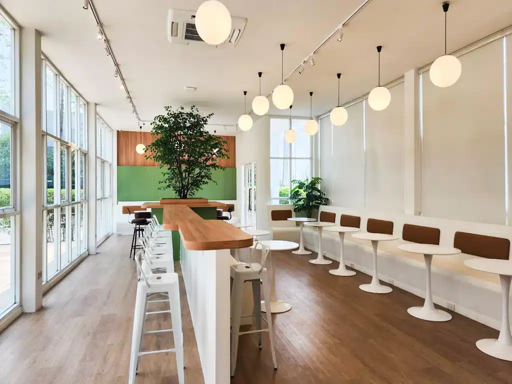 การตกแต่งภายในคาเฟ่สมัยใหม่ด้วยผนังสีขาว โต๊ะกลม เก้าอี้สีขาว พื้นไม้ หน้าต่างบานใหญ่ และโคมไฟแขวนทรงลูกโลก สร้างบรรยากาศผ่อนคลายที่มักพบในรีสอร์ทสไตล์เชียงใหม่ที่ทันสมัย ต้นไม้กระถางขนาดใหญ่ตั้งอยู่ใกล้เคาน์เตอร์.