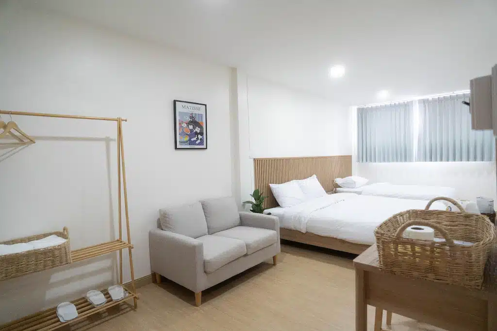 ห้องนอนที่สว่างและเรียบง่ายที่ โรงแรมน่าน ประกอบด้วยเตียงสองเตียง โซฟาขนาดเล็ก ราวไม้สำหรับแขวนเสื้อผ้า โต๊ะทำงานพร้อมตะกร้าหวาย และหน้าต่างพร้อมม่านบังแสง.