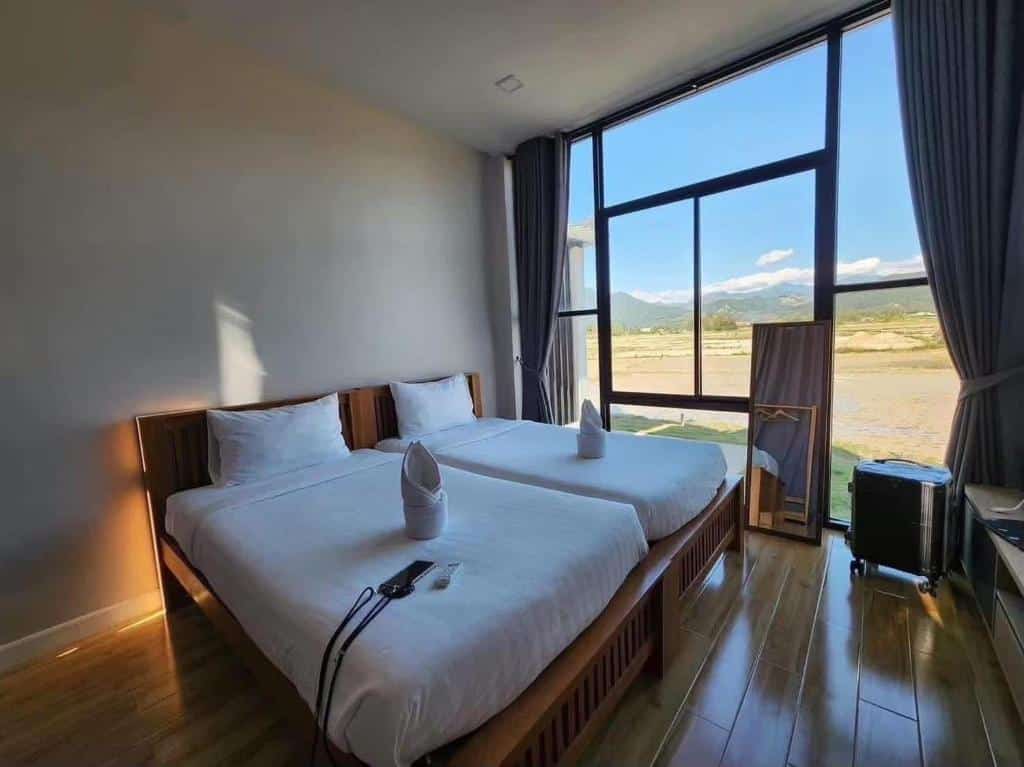 เตียงเดี่ยวสองเตียงพร้อมผ้าปูที่นอนสีขาวและตุ๊กตาผ้าขนหนูในห้องโรงแรมที่สว่างสดใส ณ รีสอร์ท น่าน พร้อมหน้าต่างบานใหญ่ที่มองเห็นทุ่งนาและภูเขาที่สวยงาม.