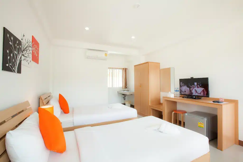 ห้องพักโรงแรมเชียงราย พร้อมเตียงเดี่ยวสองเตียง หมอนสีส้มเป็นจุดเด่น โทรทัศน์บนโต๊ะ ตู้เสื้อผ้า ตู้เย็นขนาดเล็ก และหน้าต่างที่เปิดให้แสงธรรมชาติส่องเข้ามา.